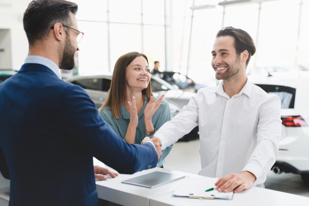 A GMC New Car Financing professional is shaking hands with the husband of a smiling happy couple who has just signed on the dotted line to buy a new vehicle