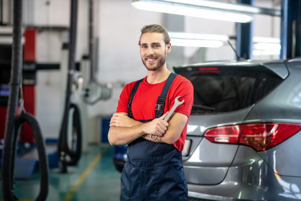 A GMC Service Center Certified Maintenance Professional in a red shirt smiles at the camera while holding a wrench with folded arms in his shop where there's a grey GMC SUV behind him.